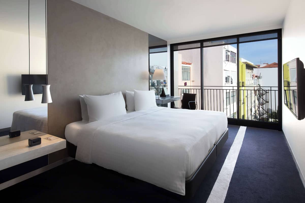 Una habitación de hotel moderna con una cama grande, mesitas de noche, un escritorio y un balcón con vistas a la ciudad.