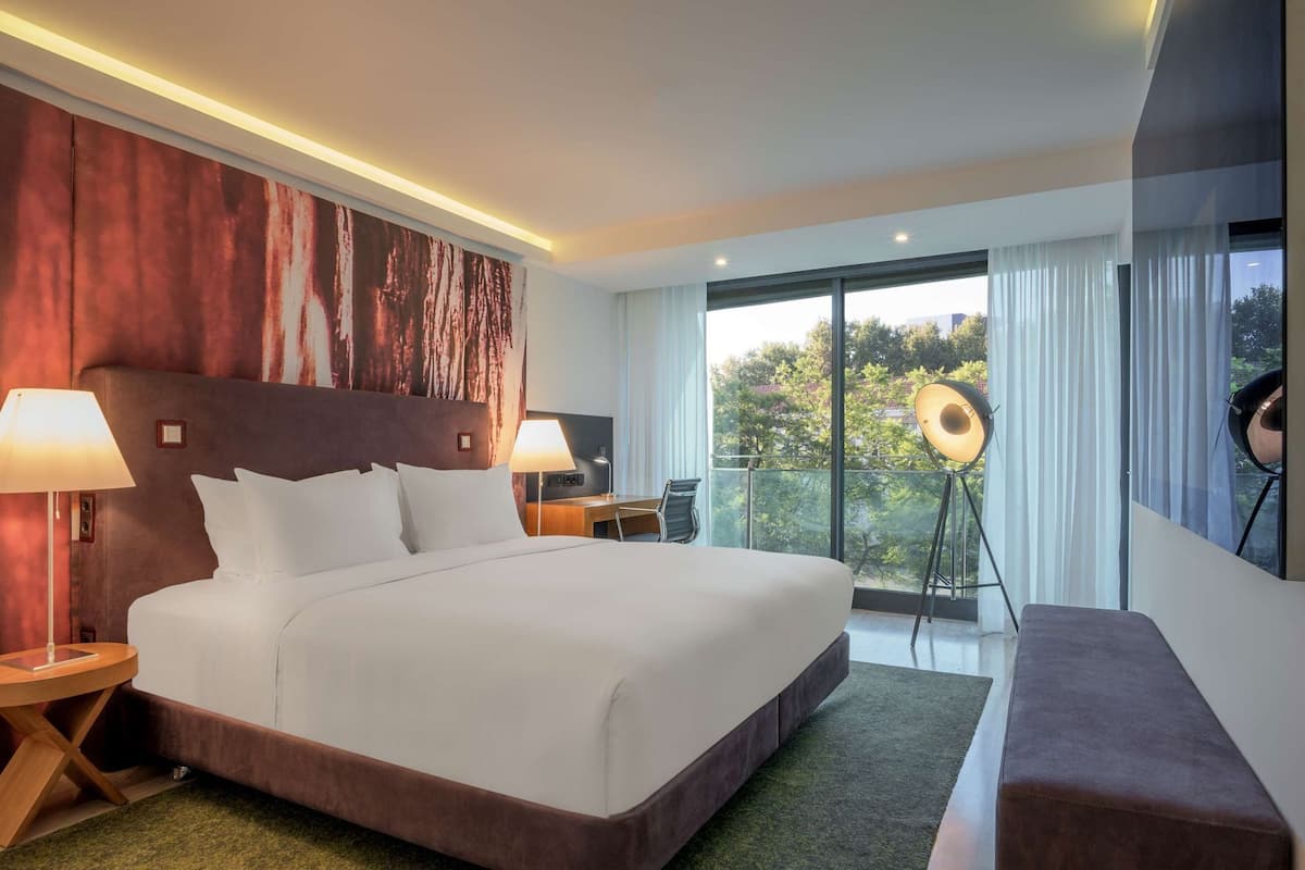 Habitación de hotel moderna con una cama grande, un escritorio, una silla, una lámpara de pie y vistas al exterior.