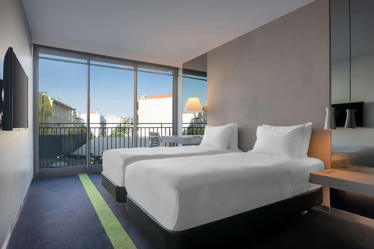 Una habitación de hotel moderna con dos camas, un balcón con mesa y sillas, y grandes ventanales con vistas a la ciudad.