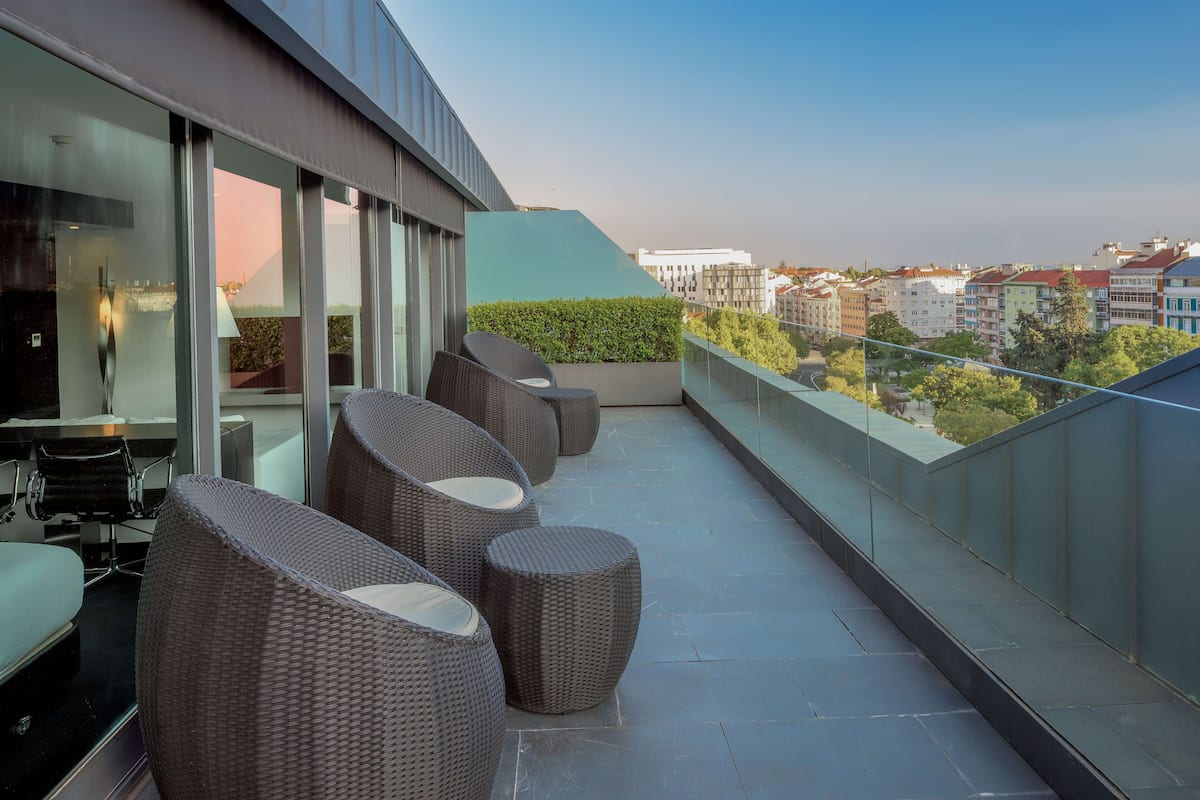 Un área de estar moderna al aire libre con mobiliario de mimbre y una barandilla de cristal con vistas a un paisaje urbano.