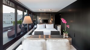 Una habitación de hotel moderna con una cama grande, una mesita de noche, un sofá con cojines, un televisor y vistas a la ciudad a través de amplios ventanales.