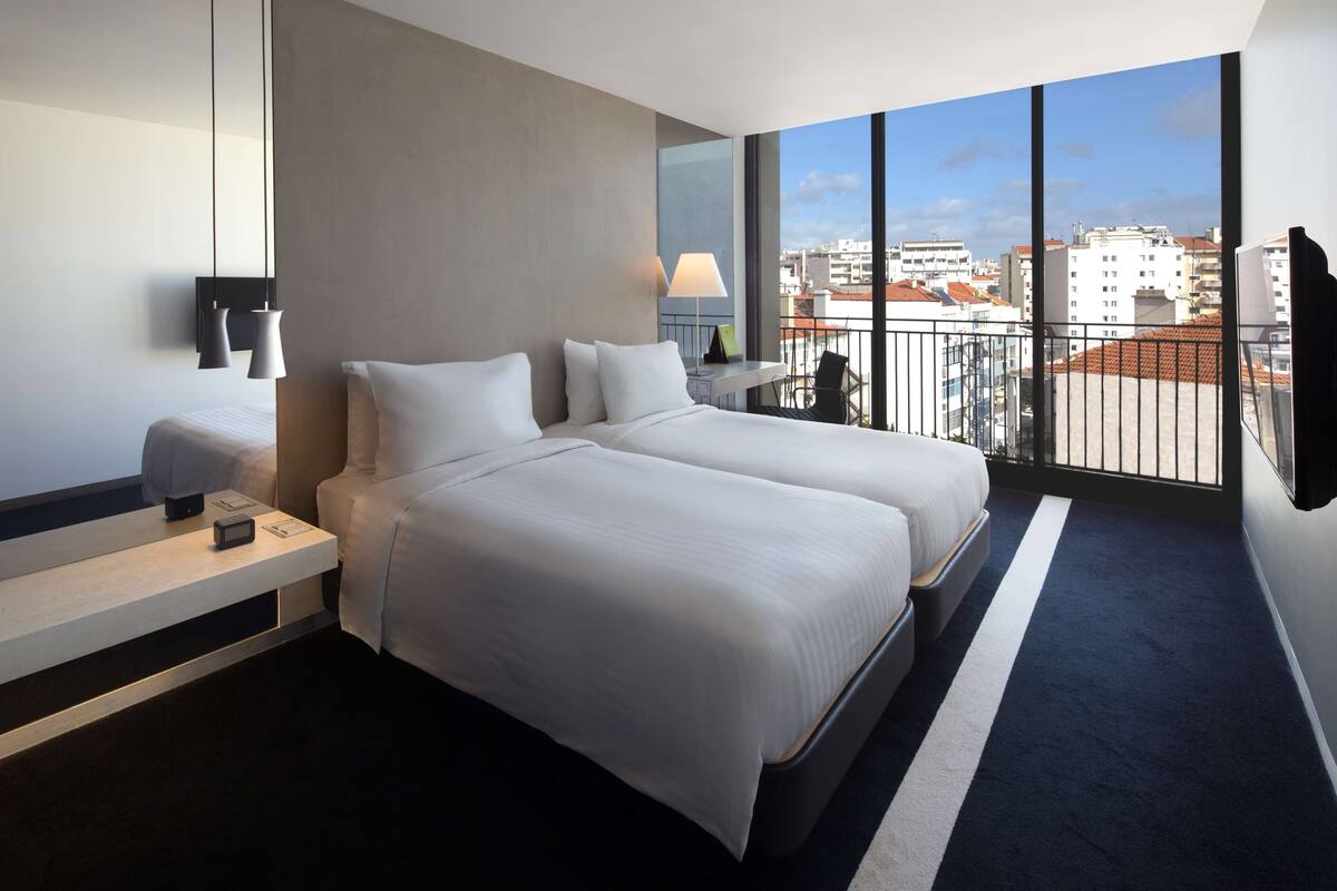 Una habitación de hotel moderna con dos camas, un ventanal amplio con vistas a la ciudad, una mesita de noche con lámpara y un escritorio pequeño con silla.