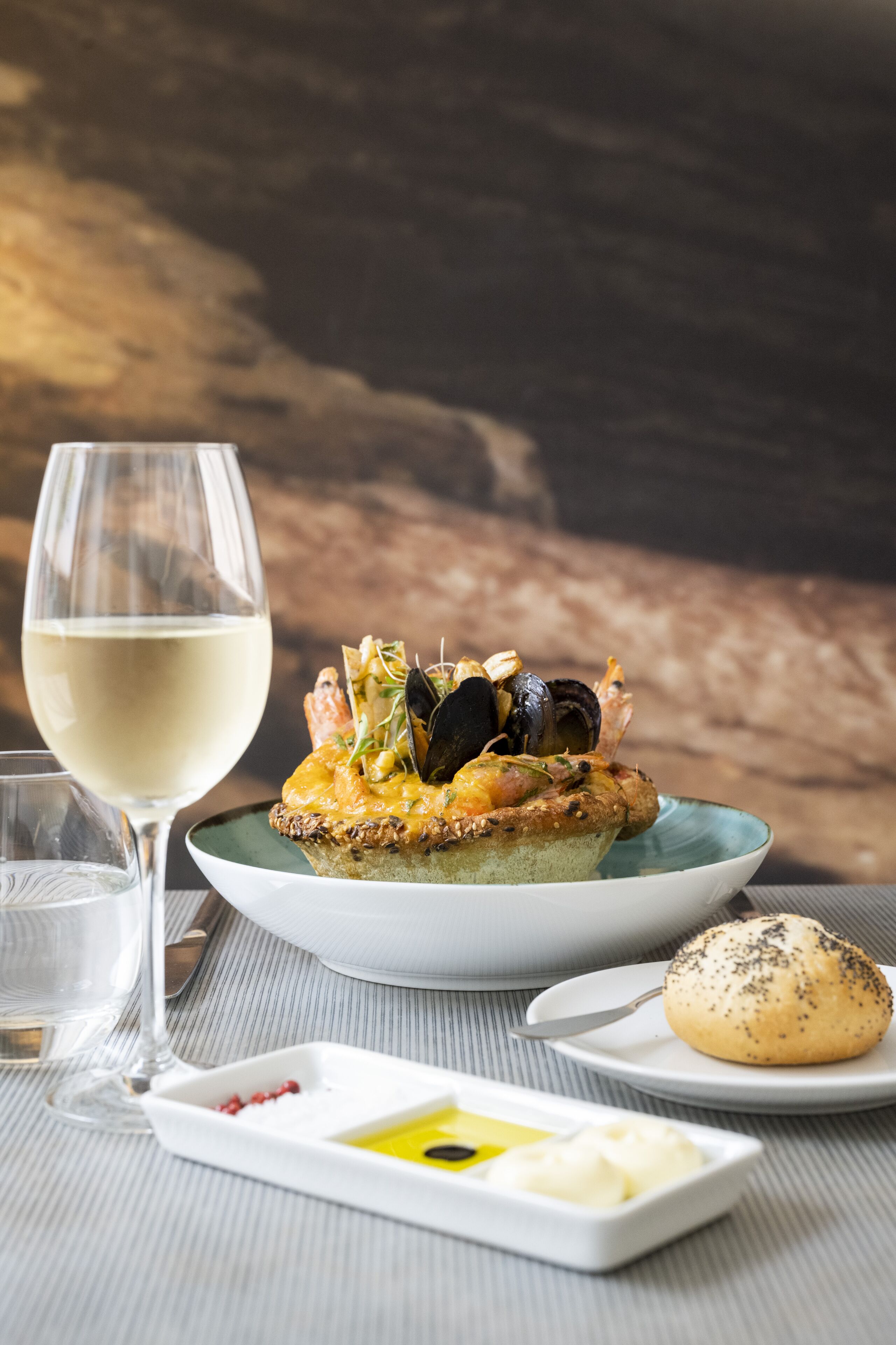 A dessert plate with a pastry topped with seafood and vegetables, a glass of white wine, and a small plate with butter and red peppercorns.