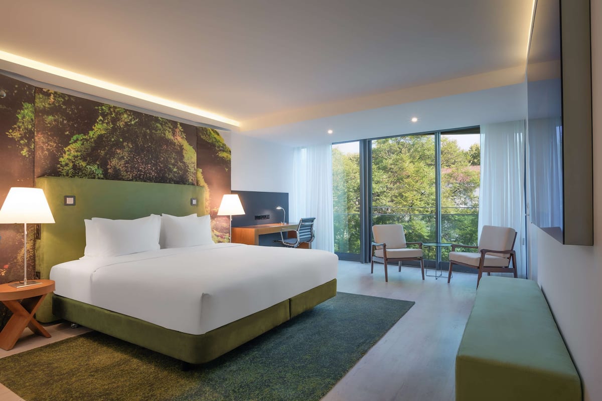 Habitación de hotel moderna con una cama grande, dos lámparas de noche, un escritorio y una silla. Se ve un ventanal con vistas a árboles.