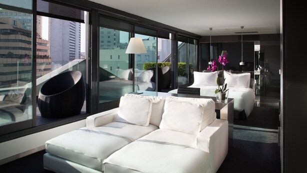 Un hotel moderno con un sofá blanco, una mesita con una lámpara y vistas a la ciudad a través de amplios ventanales.