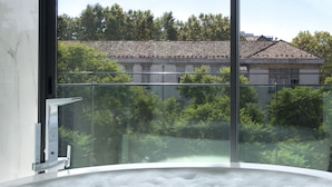 A modern bathroom with a large bathtub filled with bubbles, a glass-enclosed shower area, and a view of a residential area with trees and buildings.