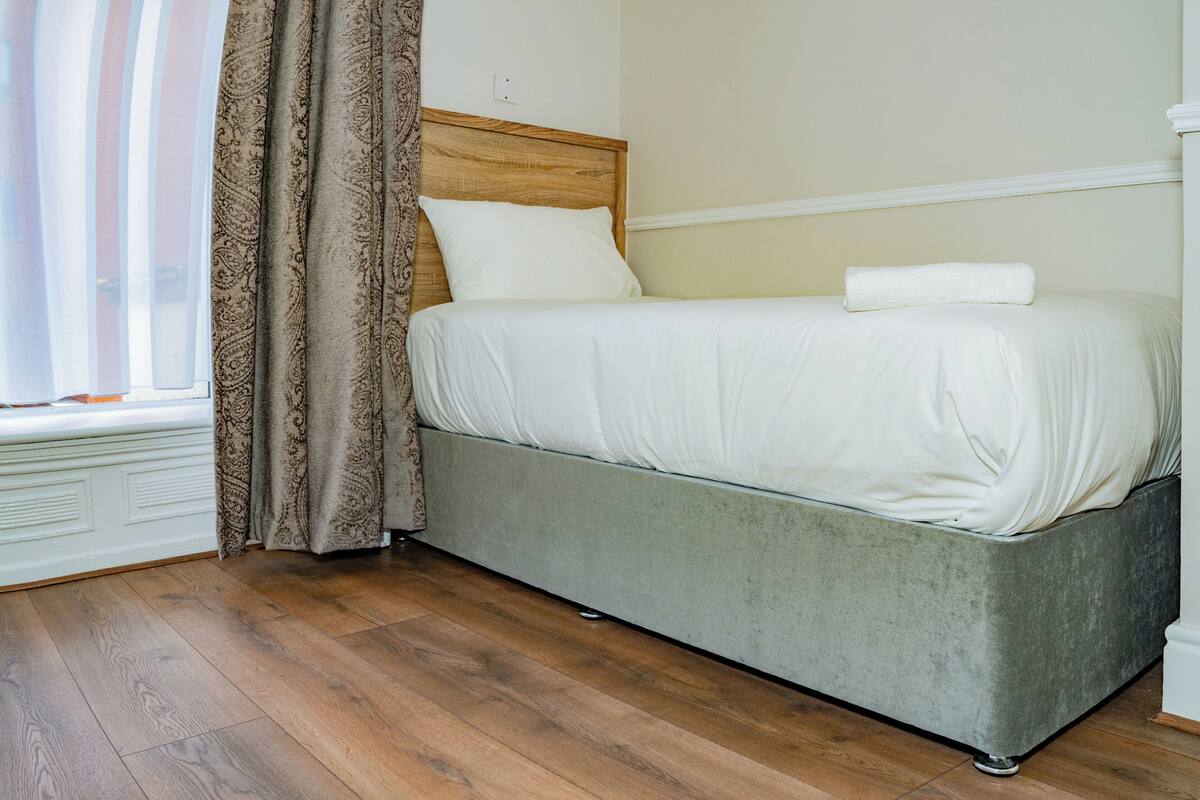 A single bed with a wooden headboard, a window with sheer curtains, and a wooden floor.