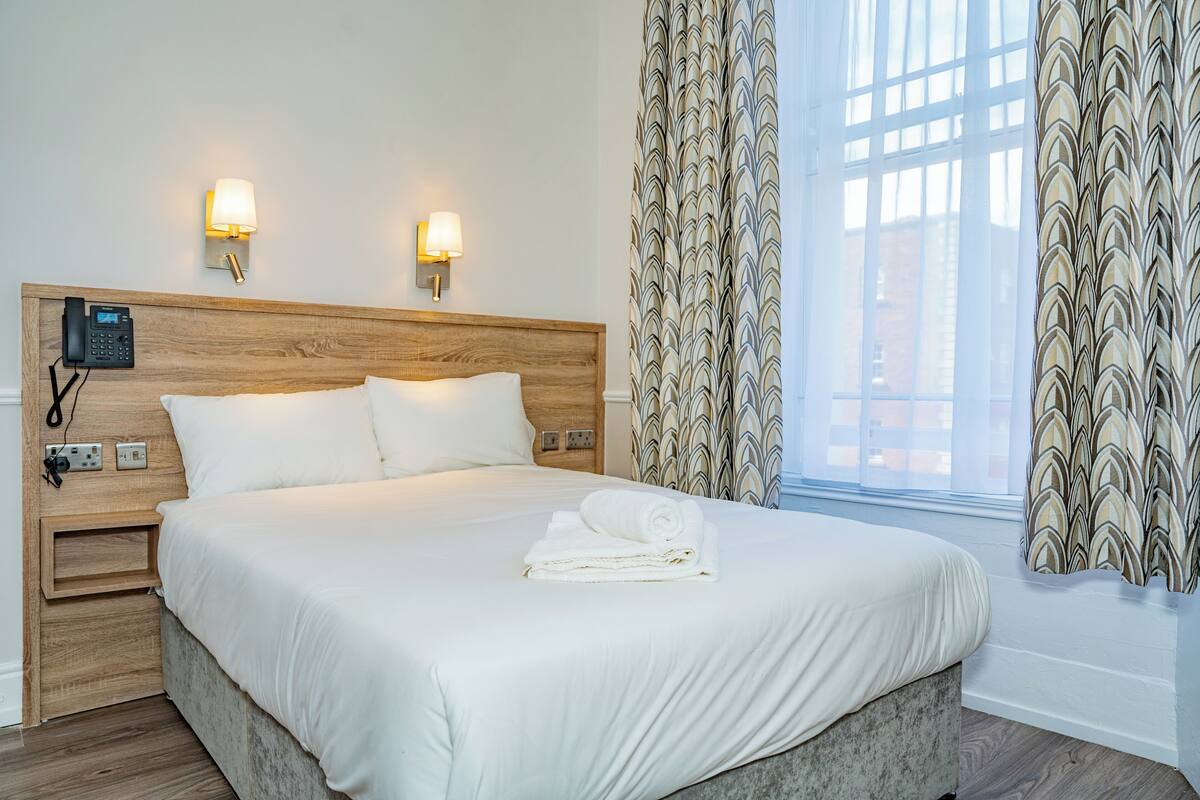 A hotel room with a wooden headboard, a bedside phone, two wall-mounted lamps, a neatly made bed with white linens, and a window with patterned curtains.