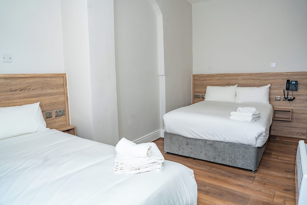 A hotel room with two beds, a grey upholstered bed, a wooden headboard, a bedside telephone, and a folded white towel on the floor.