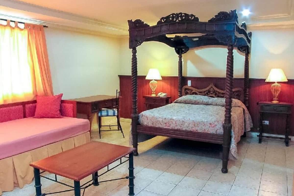 A four-poster bed with a wooden headboard, a single bed with a pink bedspread, a wooden bench, and two bedside tables with lamps.