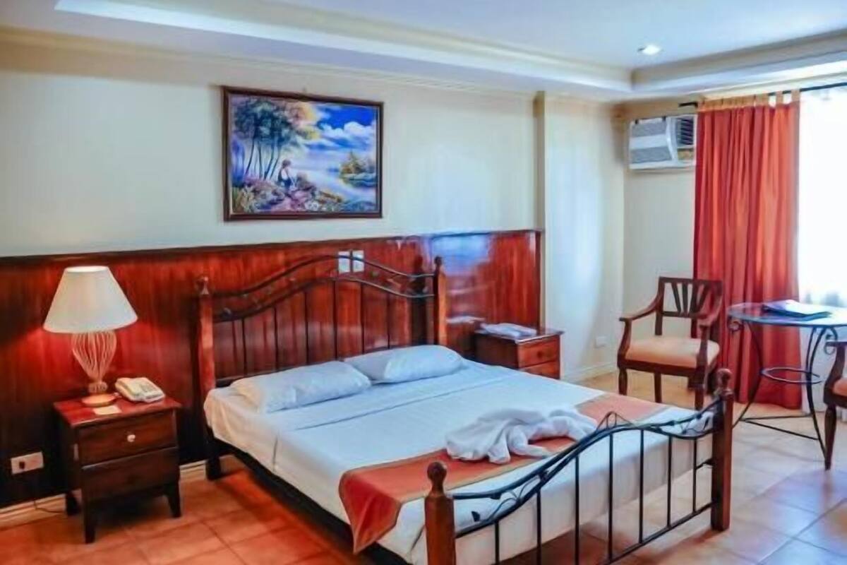 A hotel room with a bed, a nightstand, a desk, a chair, and a painting on the wall.