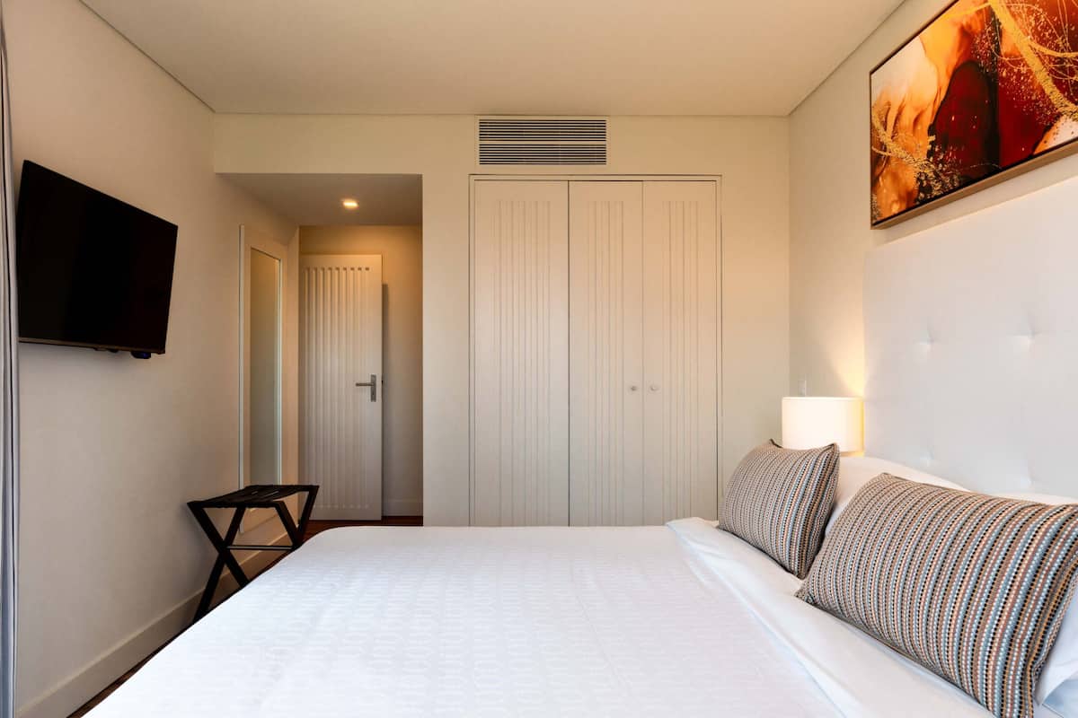Habitación de hotel con una cama grande, un televisor montado en la pared, un armario y un cuadro sobre la cama.