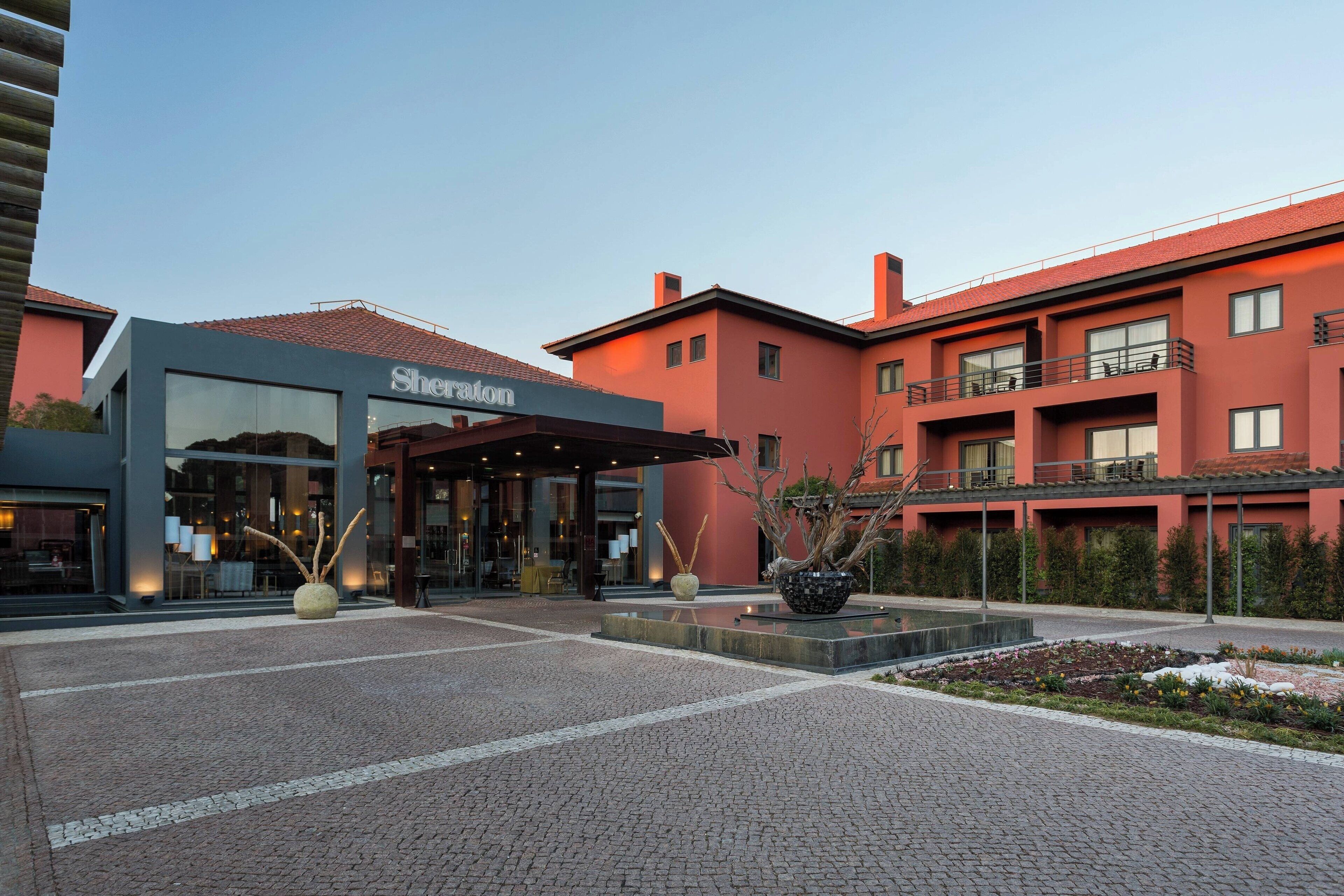 Una entrada de hotel con fachada moderna, zona de acceso cubierta y patio ajardinado con macetas decorativas.