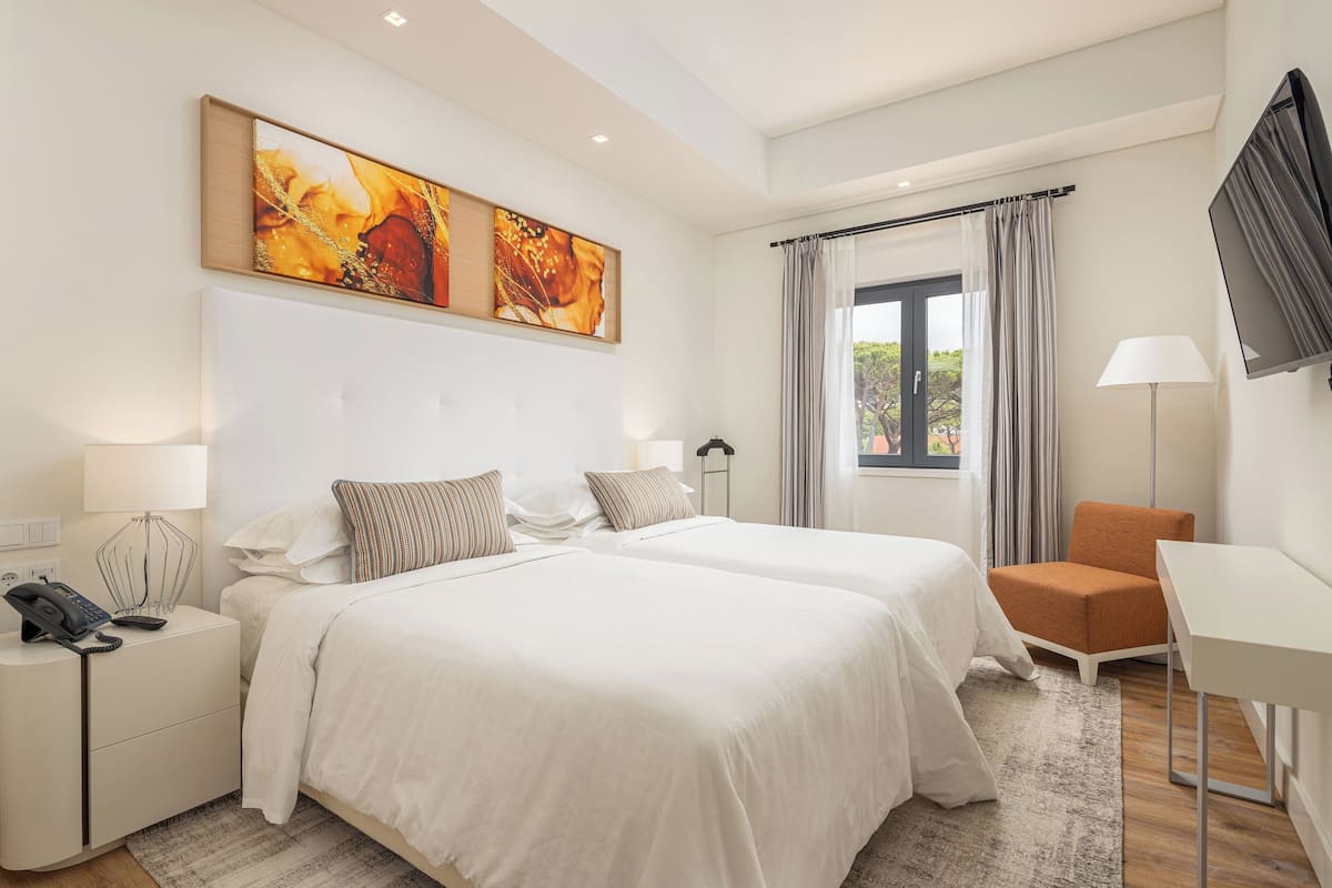 Habitación de hotel con dos camas, un escritorio, una silla, un televisor y obras de arte en la pared.
