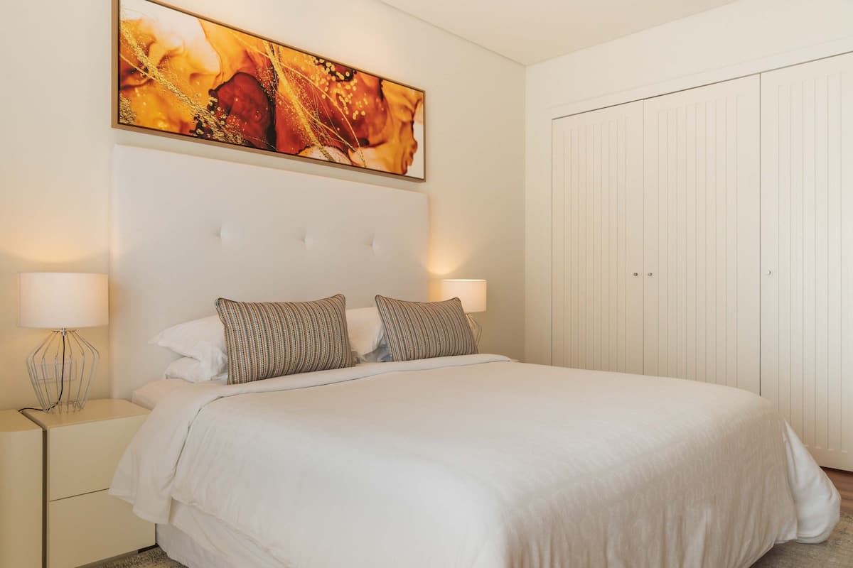 Una cama bien hecha con ropa blanca y almohadones a rayas, un cuadro abstracto grande encima y un armario blanco a la derecha.