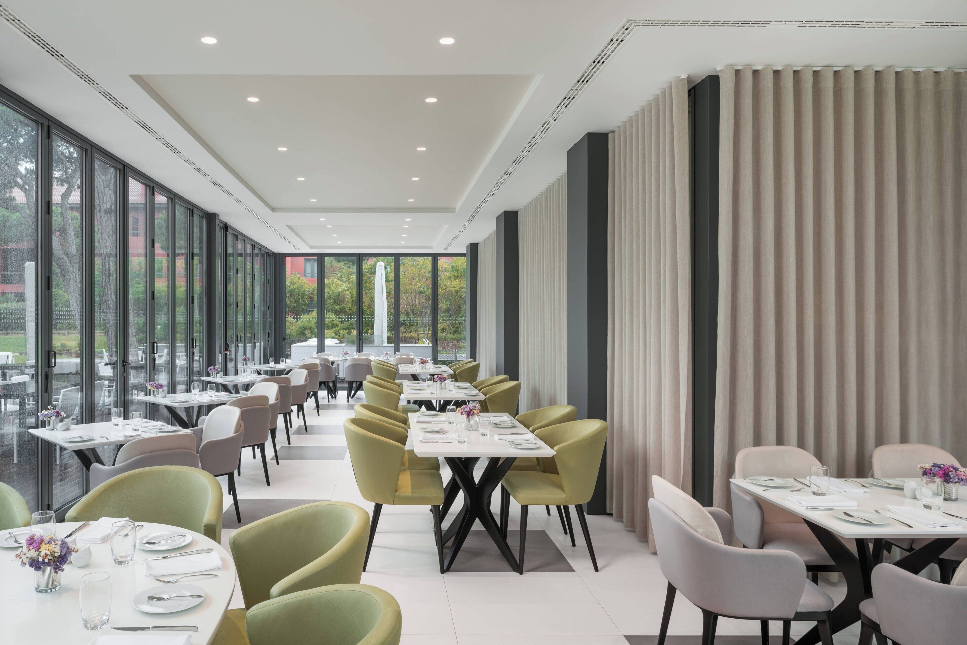 Een moderne eetruimte met tafels en stoelen, grote ramen en uitzicht op groen.