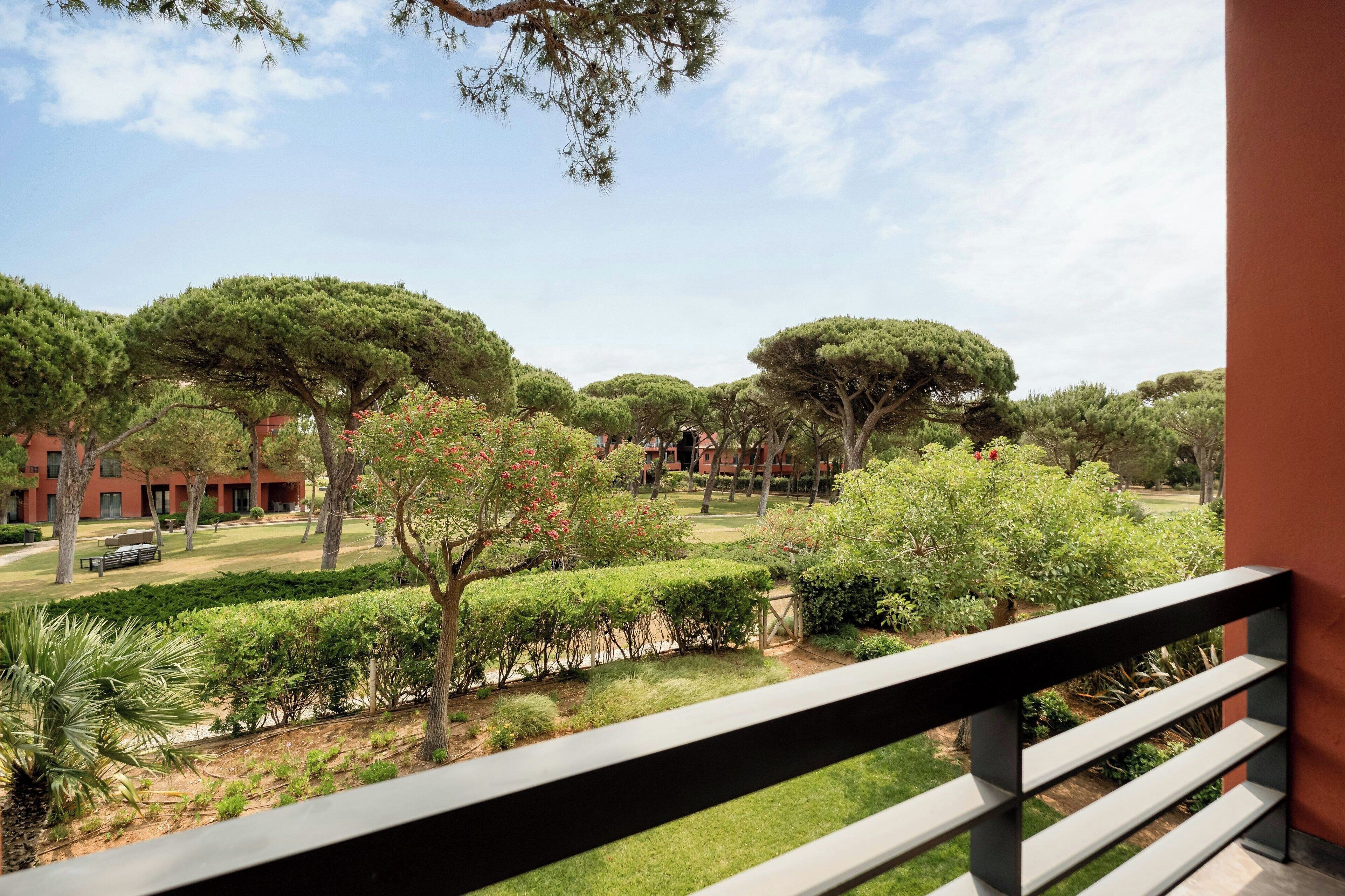 A balcony with a view of a well-maintained garden, including trees and shrubs, and a building with red brick walls in the background.
