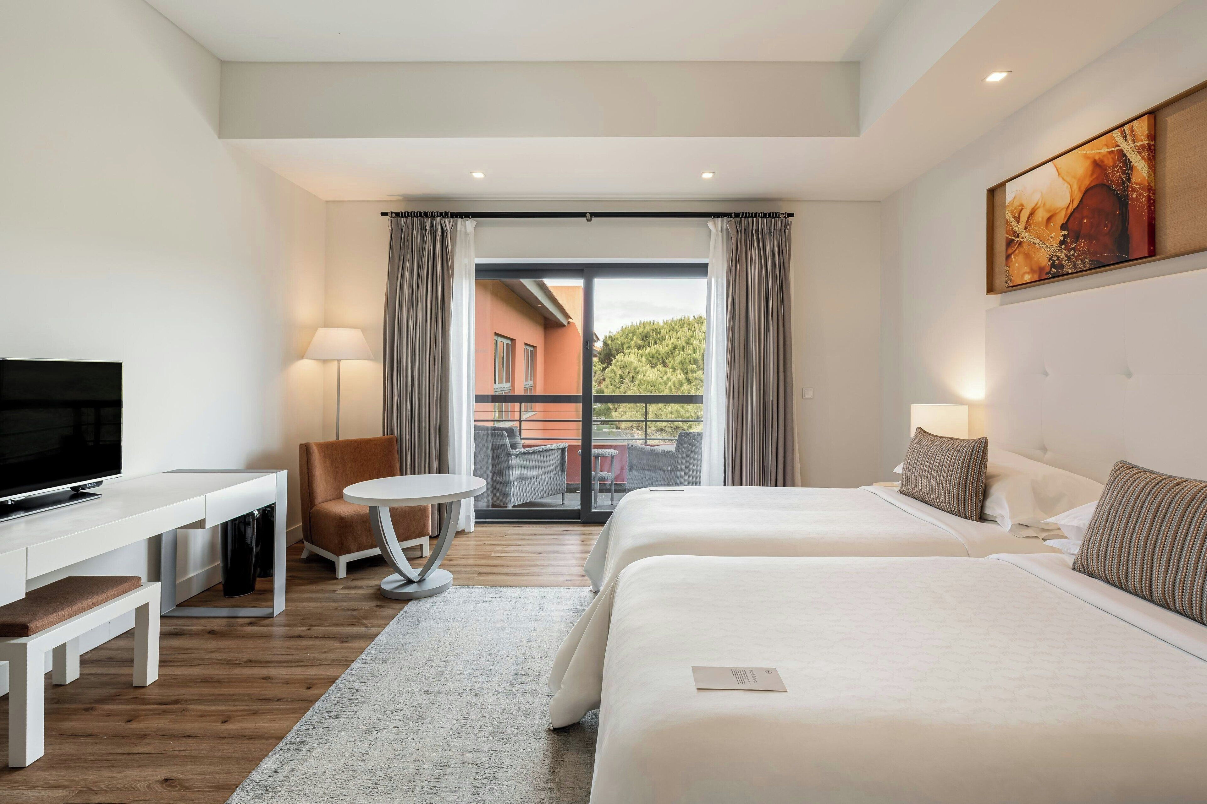 Quarto de hotel com duas camas, uma escrivaninha, uma cadeira, televisão e varanda com vista para edifícios e árvores.