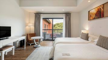 Quarto de hotel com duas camas, uma escrivaninha, uma cadeira, televisão e varanda com vista para edifícios e árvores.