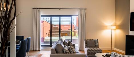 Una sala moderna con sofá, sillones, una mesita de centro y televisor. Tiene un balcón con vistas a edificios y una piscina.