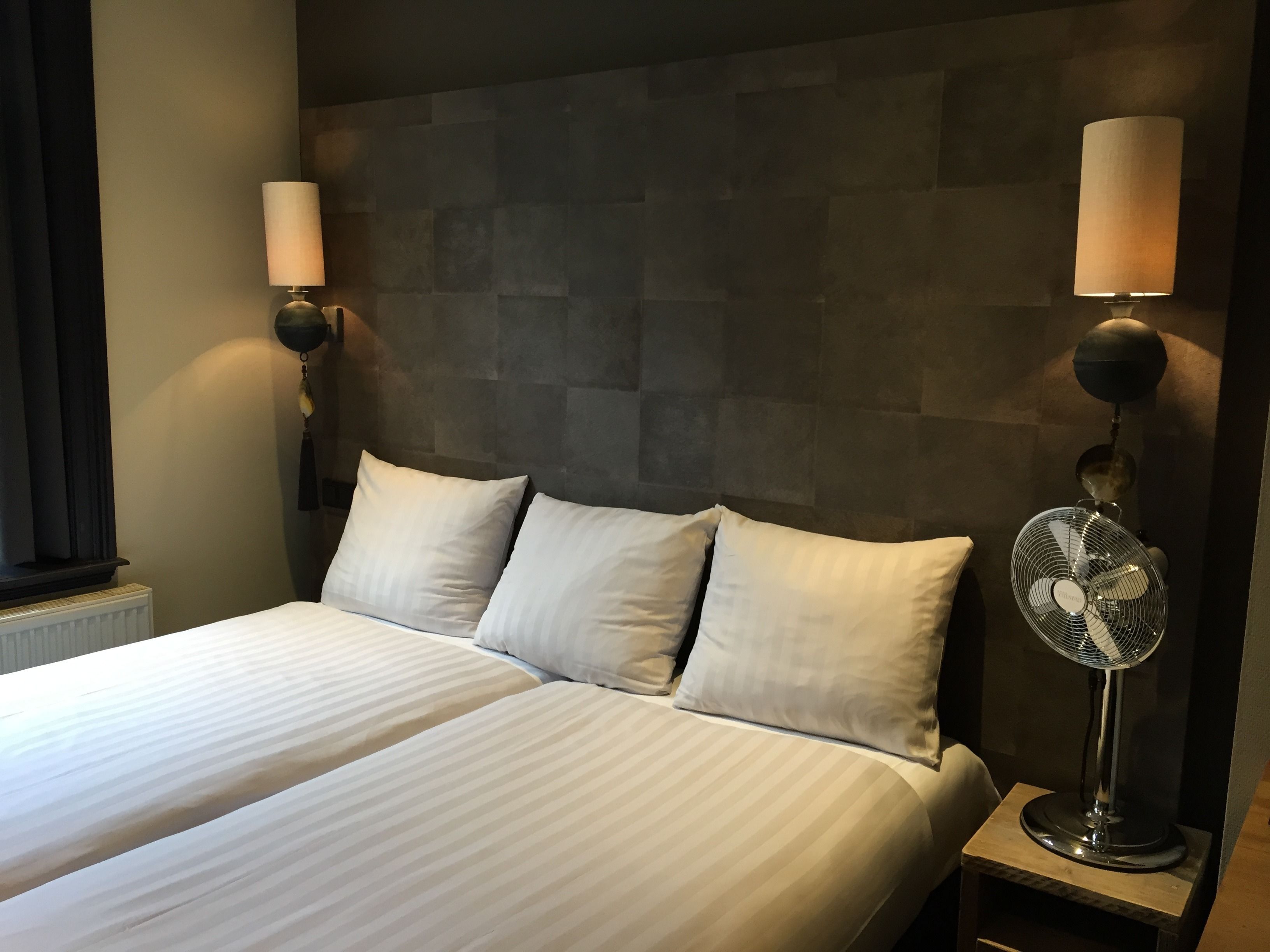 Una cama doble con ropa de cama blanca, dos lámparas fijadas a la pared y un ventilador de pie.