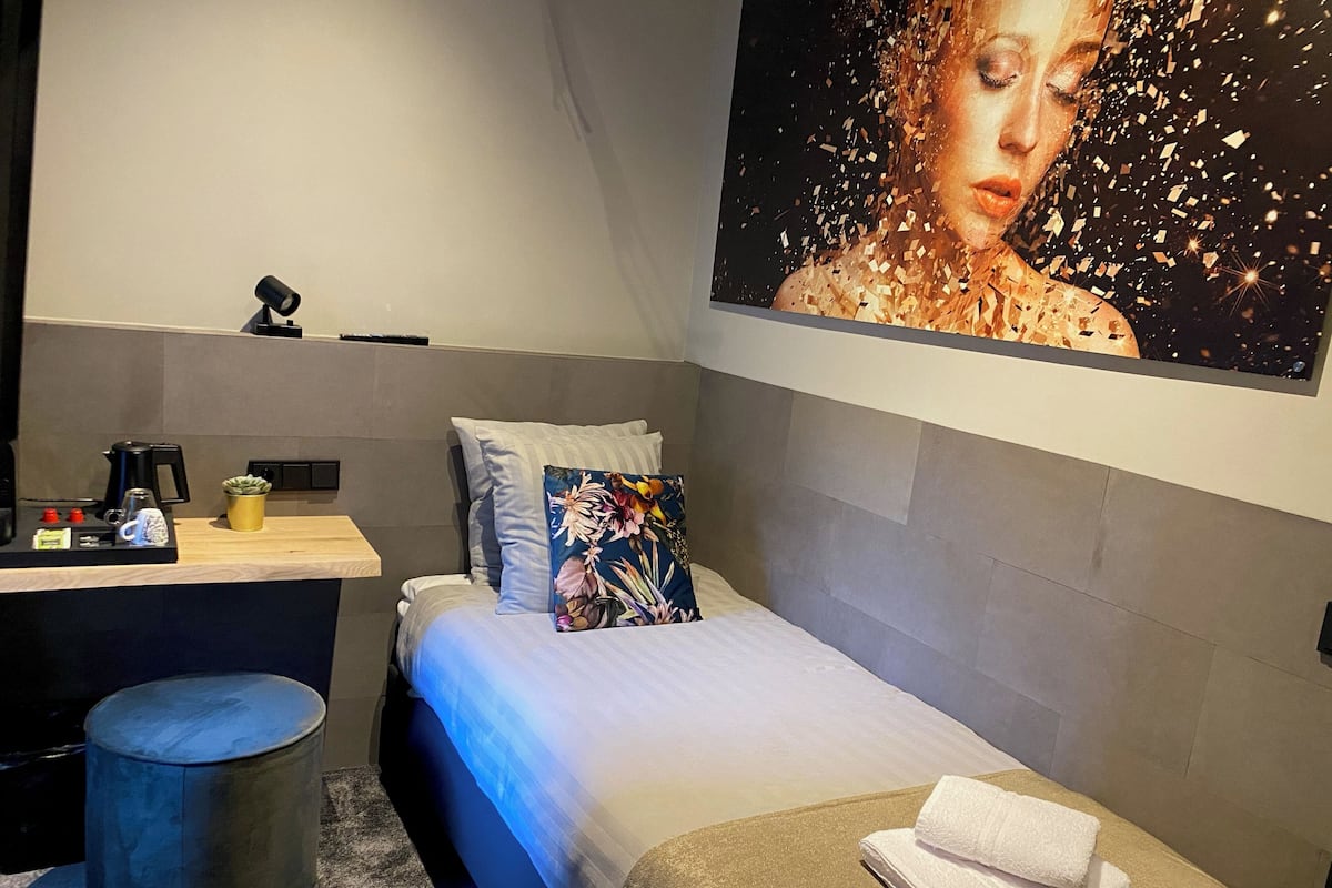 Une chambre à coucher comprenant un lit, une table de chevet avec une machine à café, un petit tabouret et une œuvre d’art accrochée au mur.