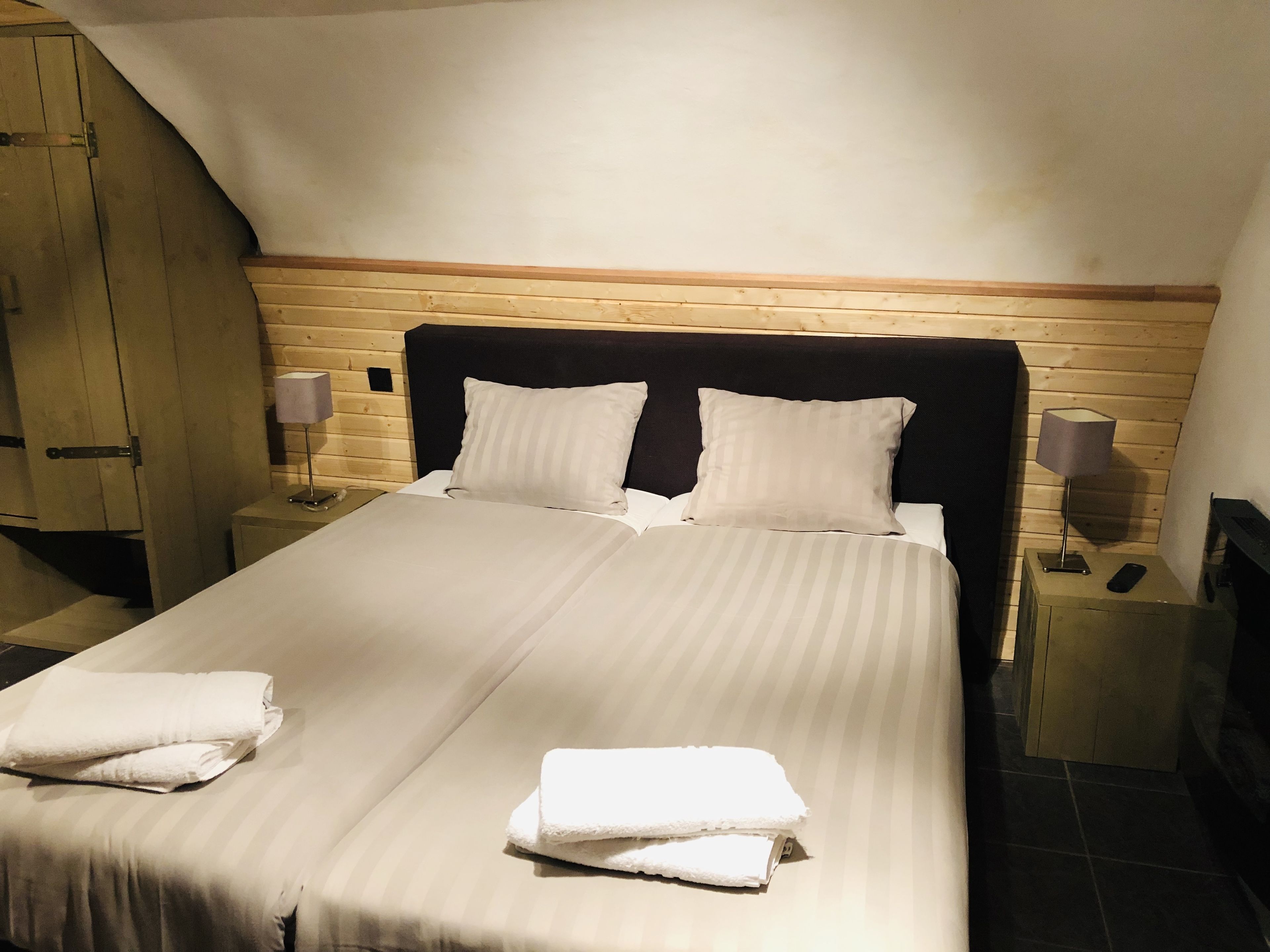 Una cama bien hecha con dos almohadas y dos toallas dobladas, cabecera de madera, dos mesitas de noche con lámparas y un armario de madera.