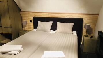 Una cama bien hecha con dos almohadas y dos toallas dobladas, cabecera de madera, dos mesitas de noche con lámparas y un armario de madera.