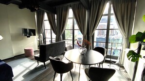Una sala de estar moderna con una mesa redonda, cuatro sillas, un sofá, un televisor y ventanas grandes con cortinas.