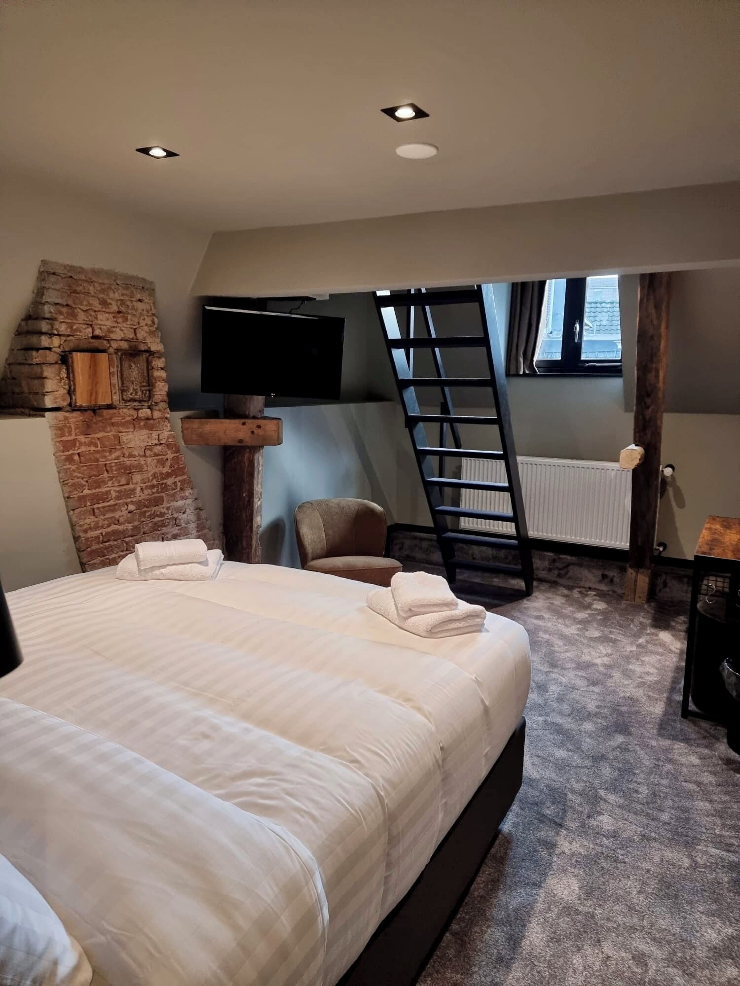 Un dormitorio con una cama, un televisor montado en una pared de ladrillo, una escalera y una ventana.