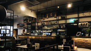 Zona de bar con mesas y sillas, estanterías con botellas y vasos, y una barra con caja registradora.