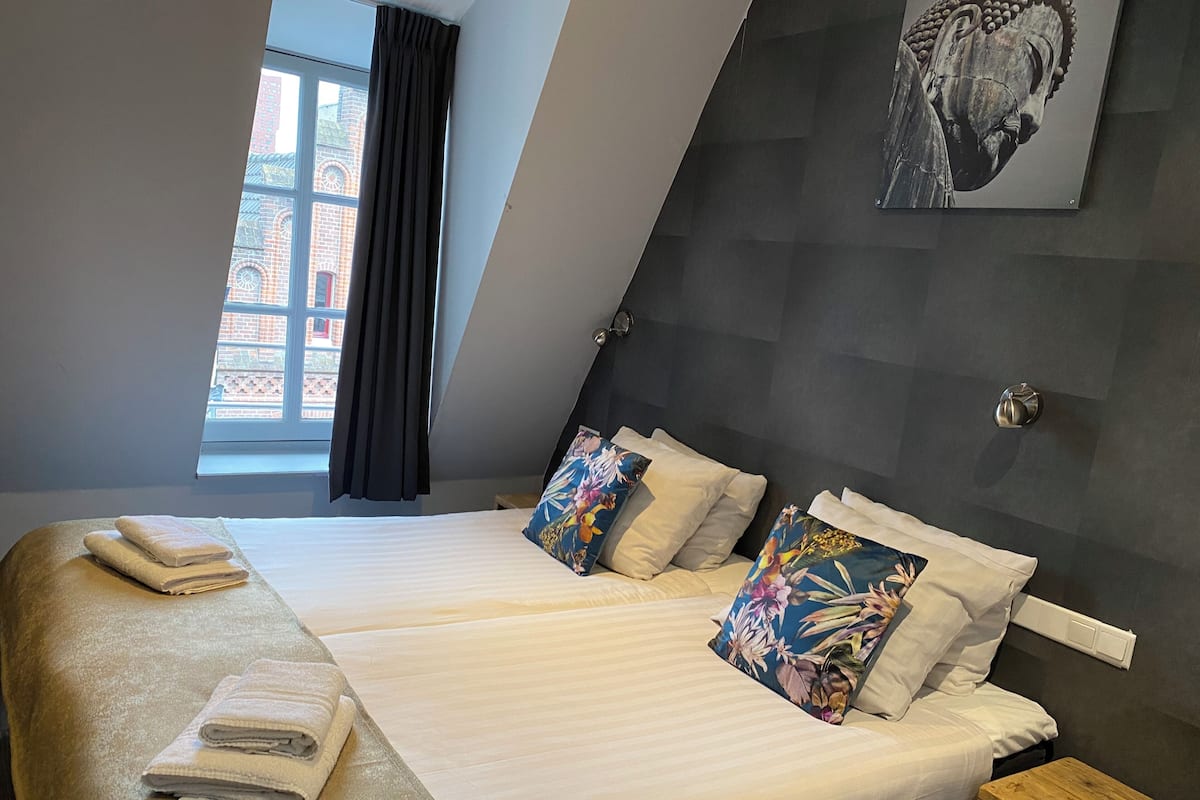 Une chambre d’hôtel comprenant un lit, des oreillers, une table de chevet, une fenêtre avec des rideaux et une œuvre d’art accrochée au mur.