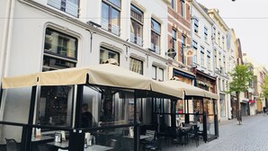 Mesas de café al aire libre con sombrillas, un cartel de Heineken y diversos comercios en una calle adoquinada.