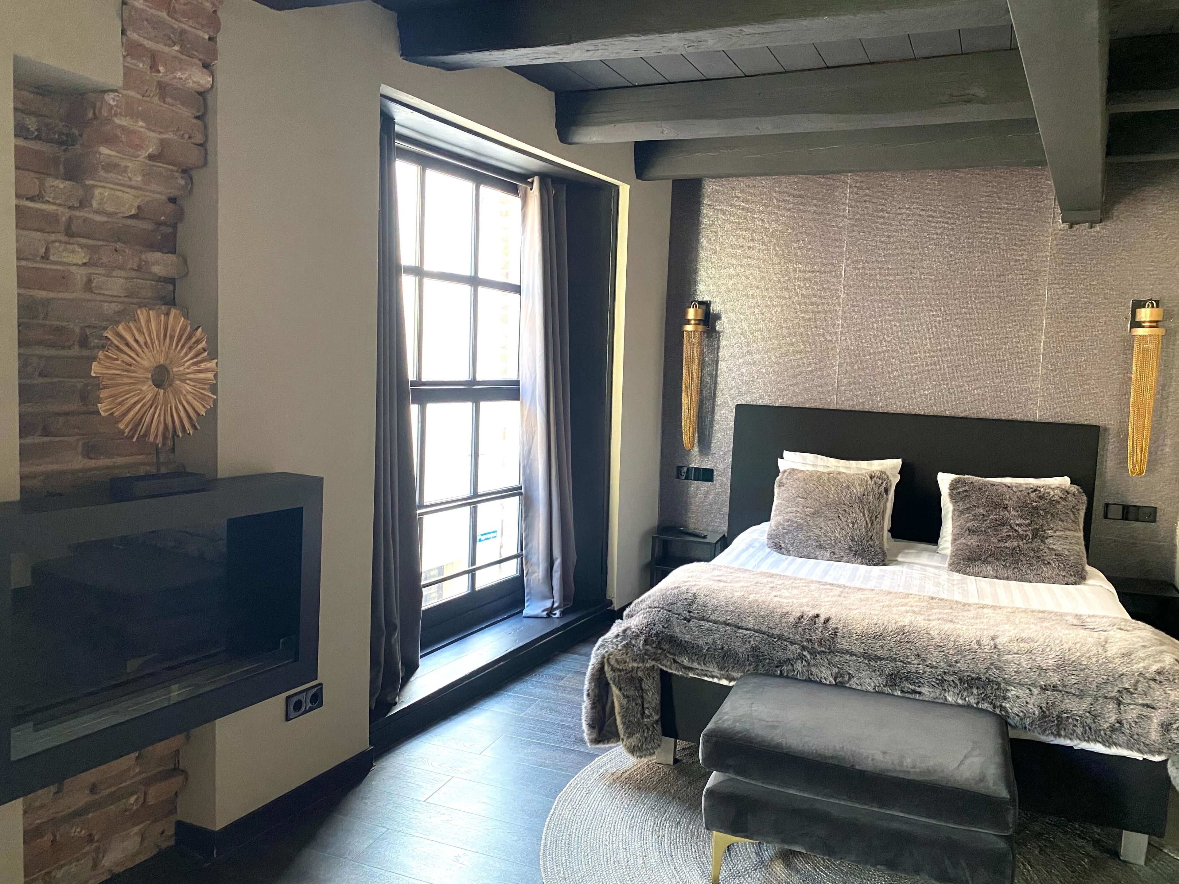 A bedroom with a bed, bench, fireplace, and large window.