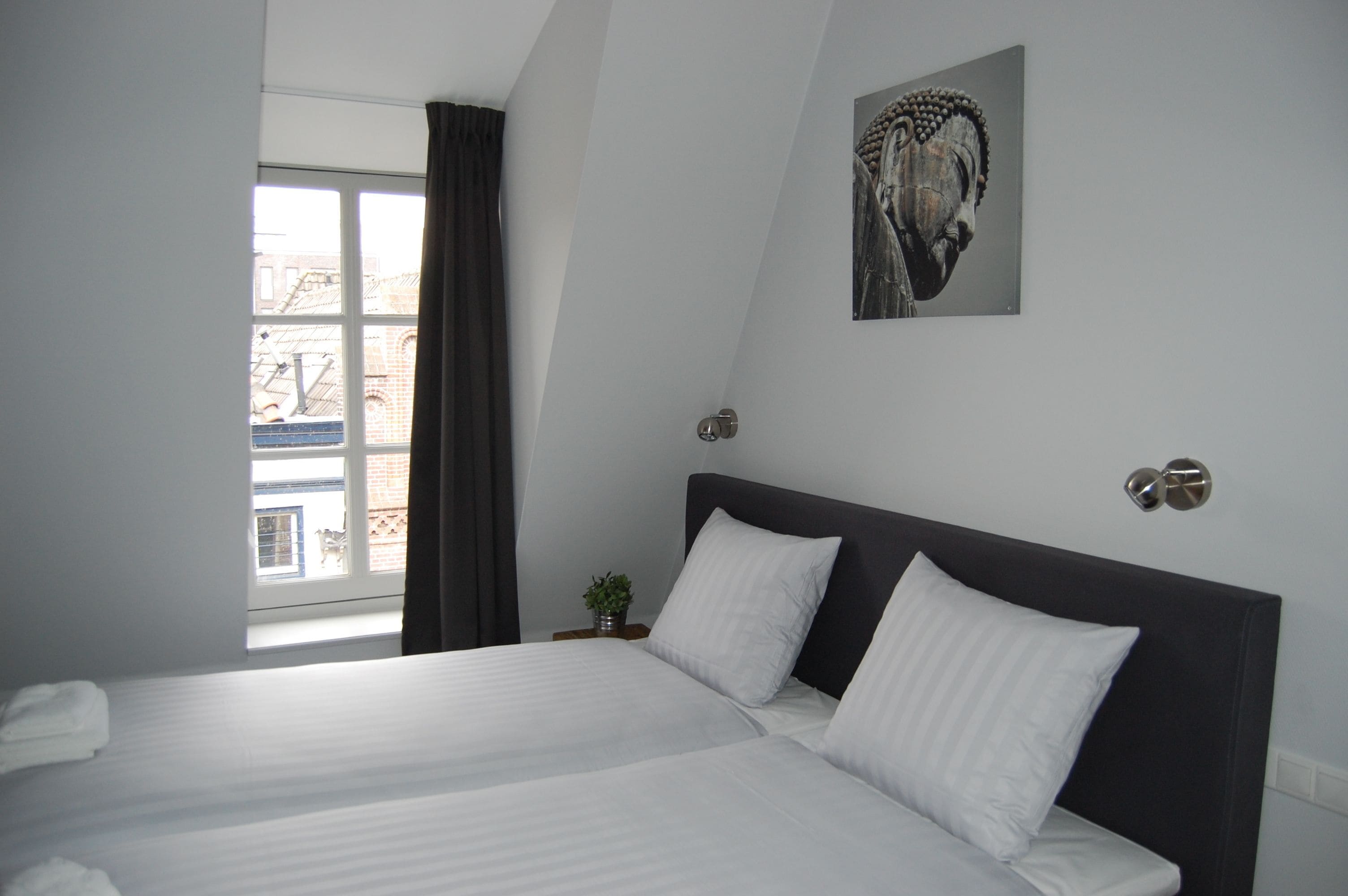 Una habitación de hotel moderna con una cama grande, una ventana con cortinas, una planta pequeña y una obra de arte colgada en la pared.