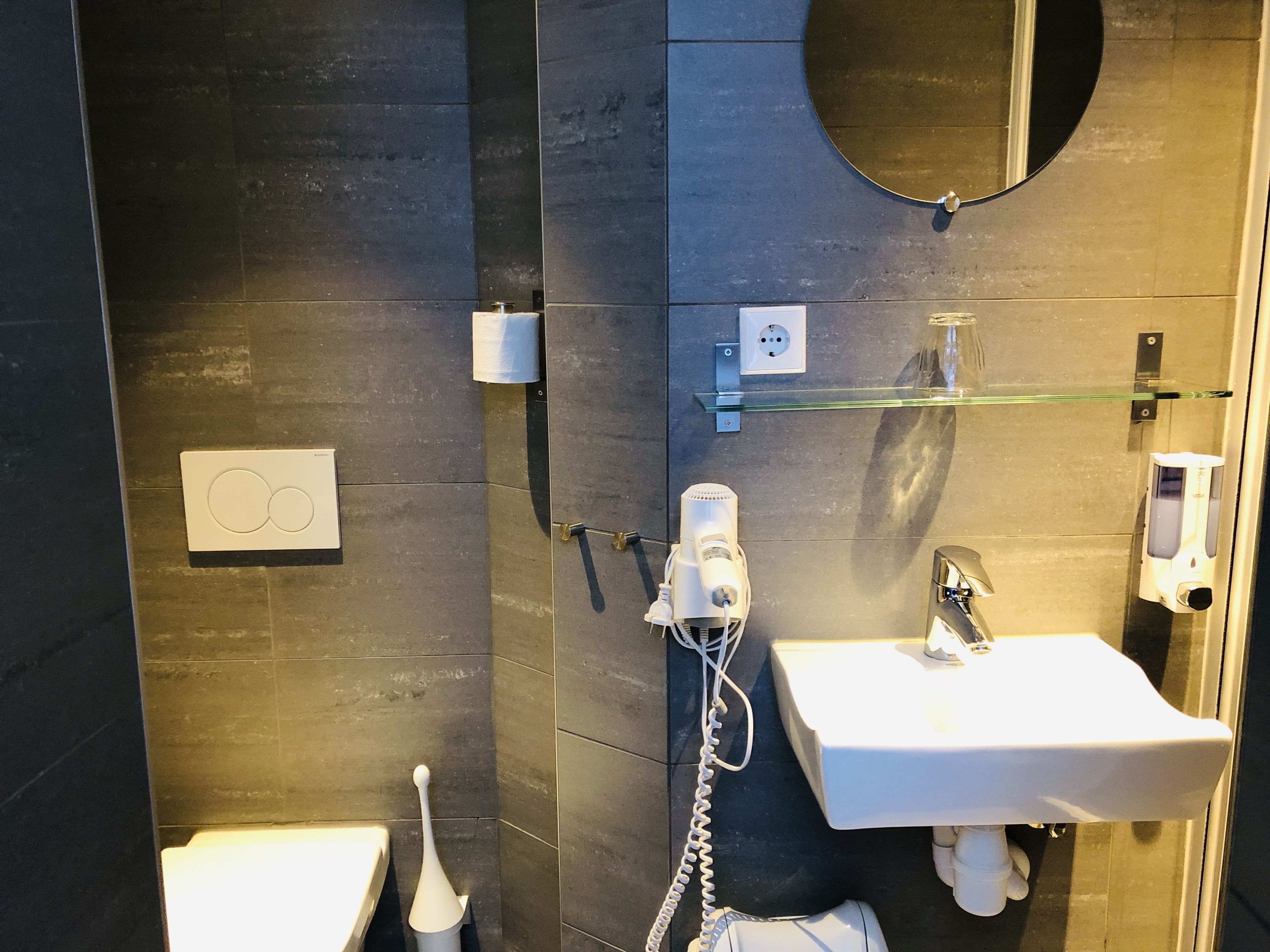 Un baño moderno con una pared de azulejos oscuros, un lavamanos blanco, un cabezal de ducha de mano, un inodoro y un estante de vidrio.