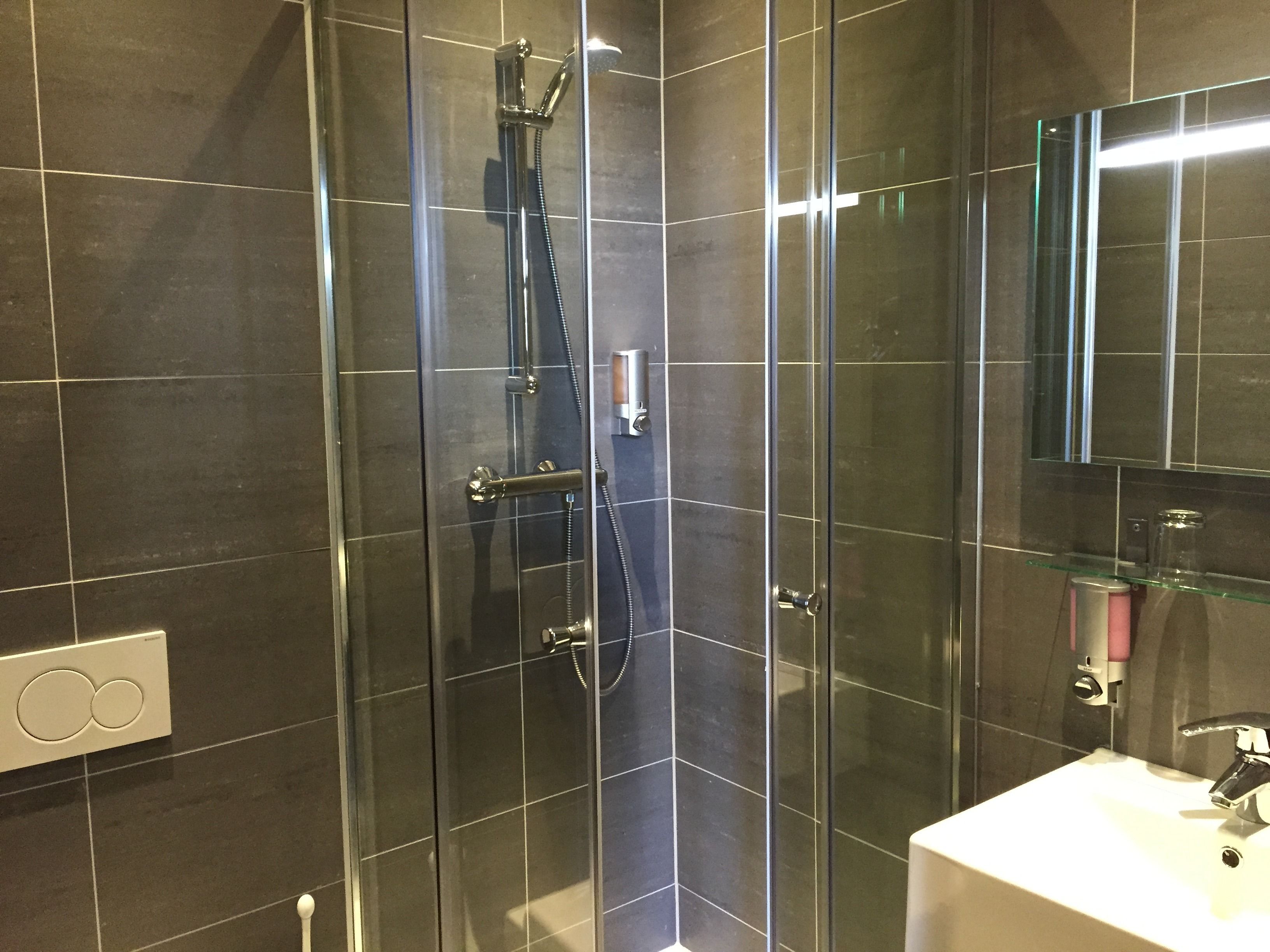 Un bagno moderno con piastrelle scure, una cabina doccia in vetro, un lavabo bianco e uno specchio sopra il lavabo.