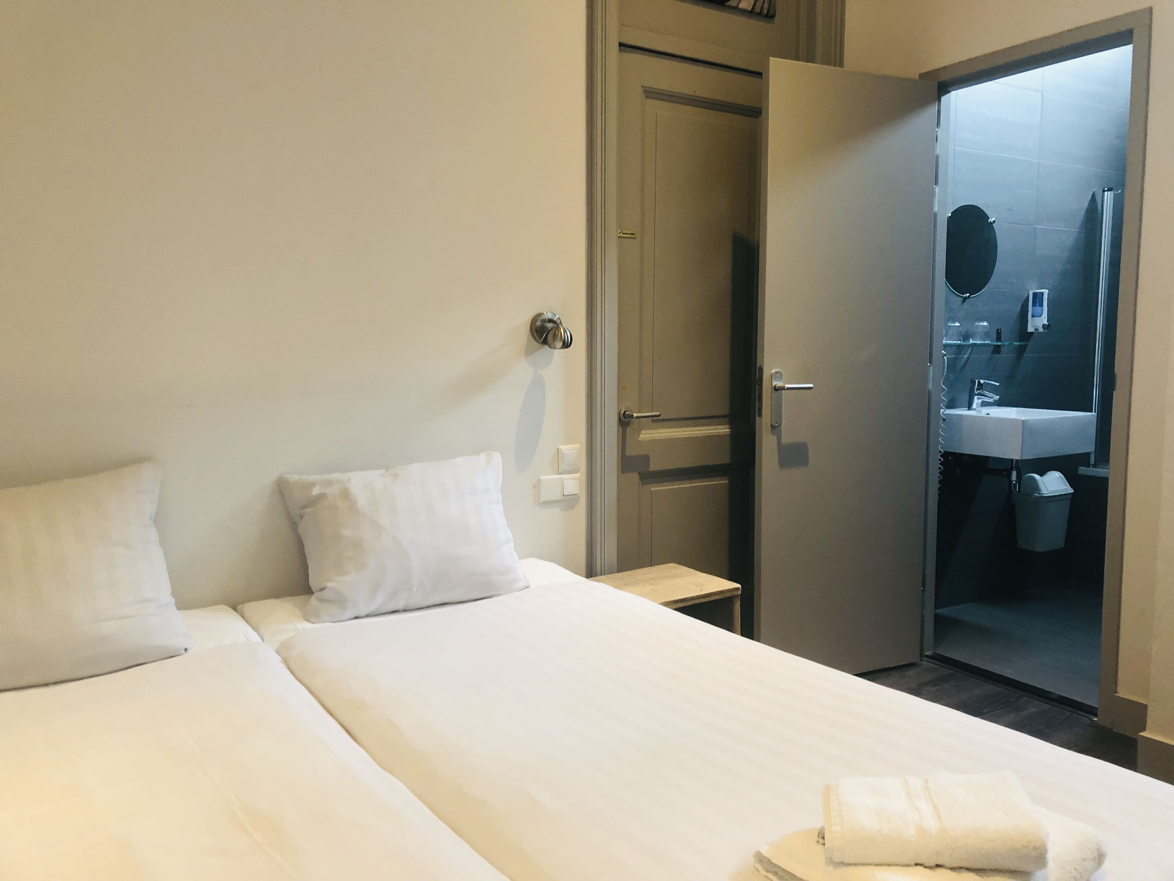 Habitación de hotel con cama, dos almohadas, mesita de noche y puerta abierta que da a un baño con lavabo y espejo.