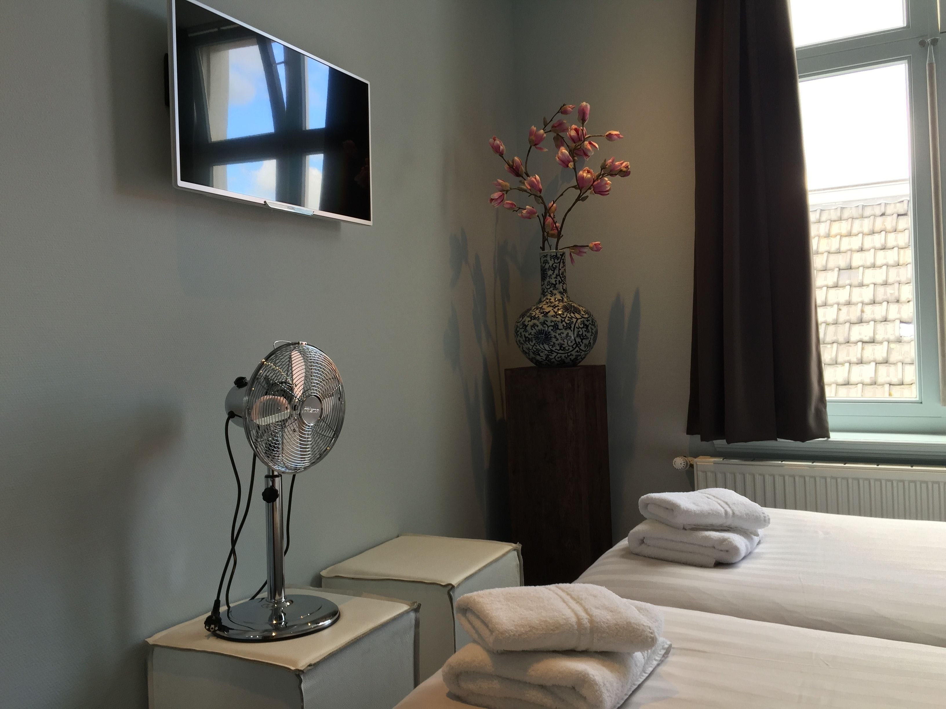 Une chambre d’hôtel dotée d’un téléviseur à écran plat fixé au mur, d’un ventilateur sur pied, d’un vase avec des fleurs, d’une fenêtre avec des rideaux et de lits soigneusement faits avec des serviettes.