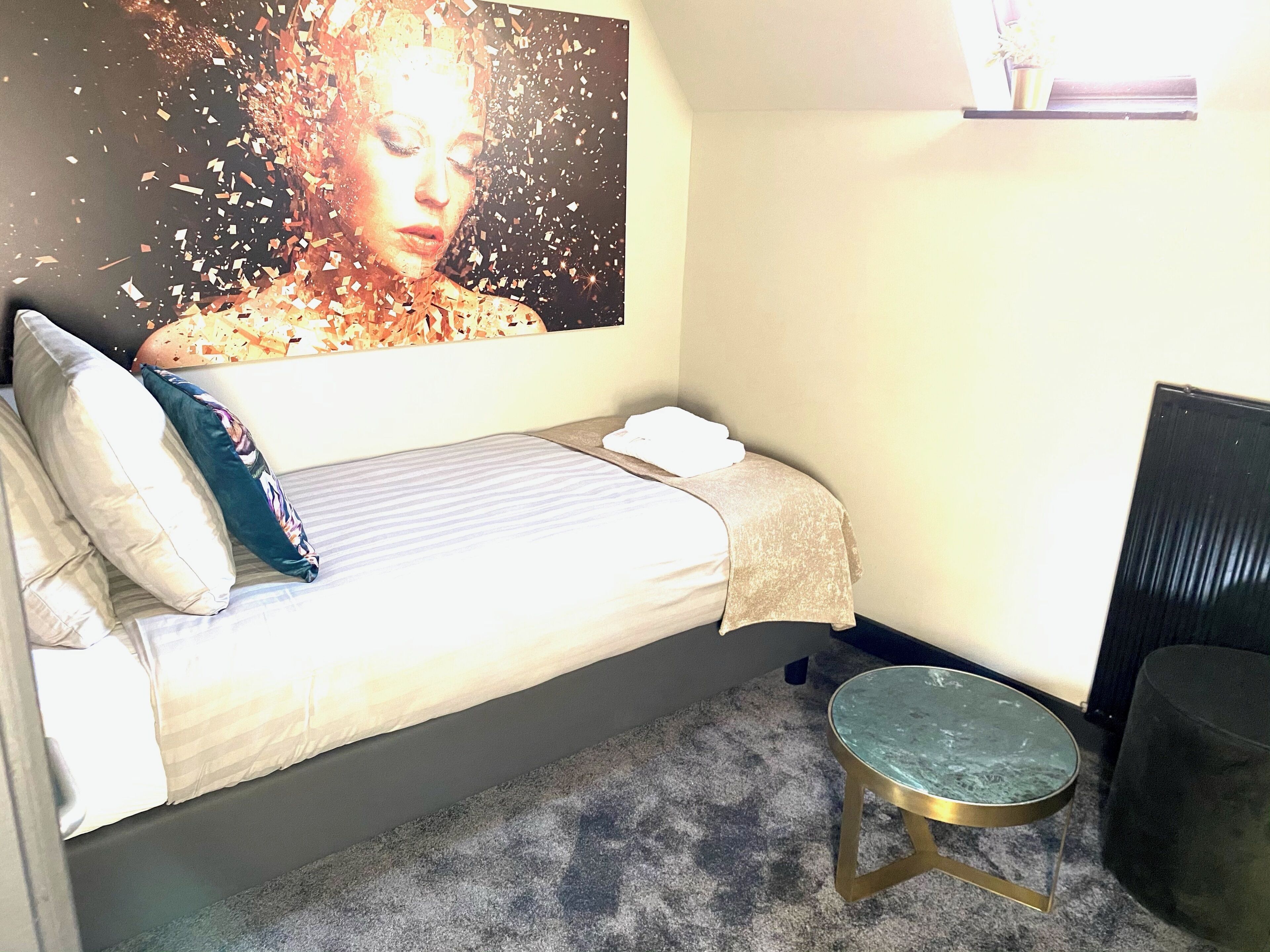 Een slaapkamer met een bed, kussens, een klein rond tafeltje en een wanddecoratie.