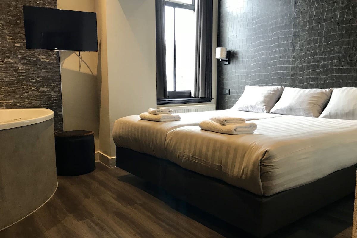 Une chambre d’hôtel moderne avec un grand lit, un téléviseur à écran plat fixé au mur, une fenêtre avec des rideaux et un mur en briques de couleur sombre.