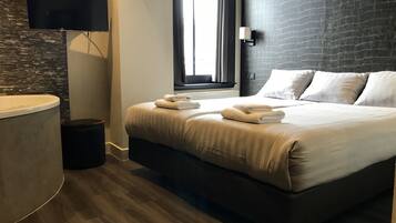 A modern hotel room with a large bed, a flat-screen TV mounted on the wall, a window with curtains, and a dark brick wall.