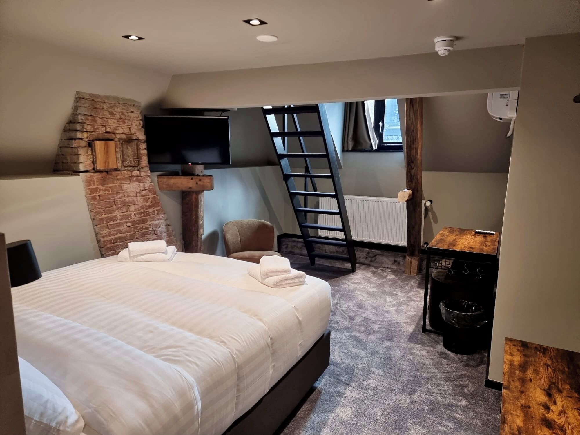 Um quarto com cama, parede de tijolos, televisão, cadeira, escada e radiador.