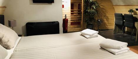 A neatly made bed with white linens and a folded towel, a television mounted on the wall, a wooden ceiling, and a small table with chairs in the background.