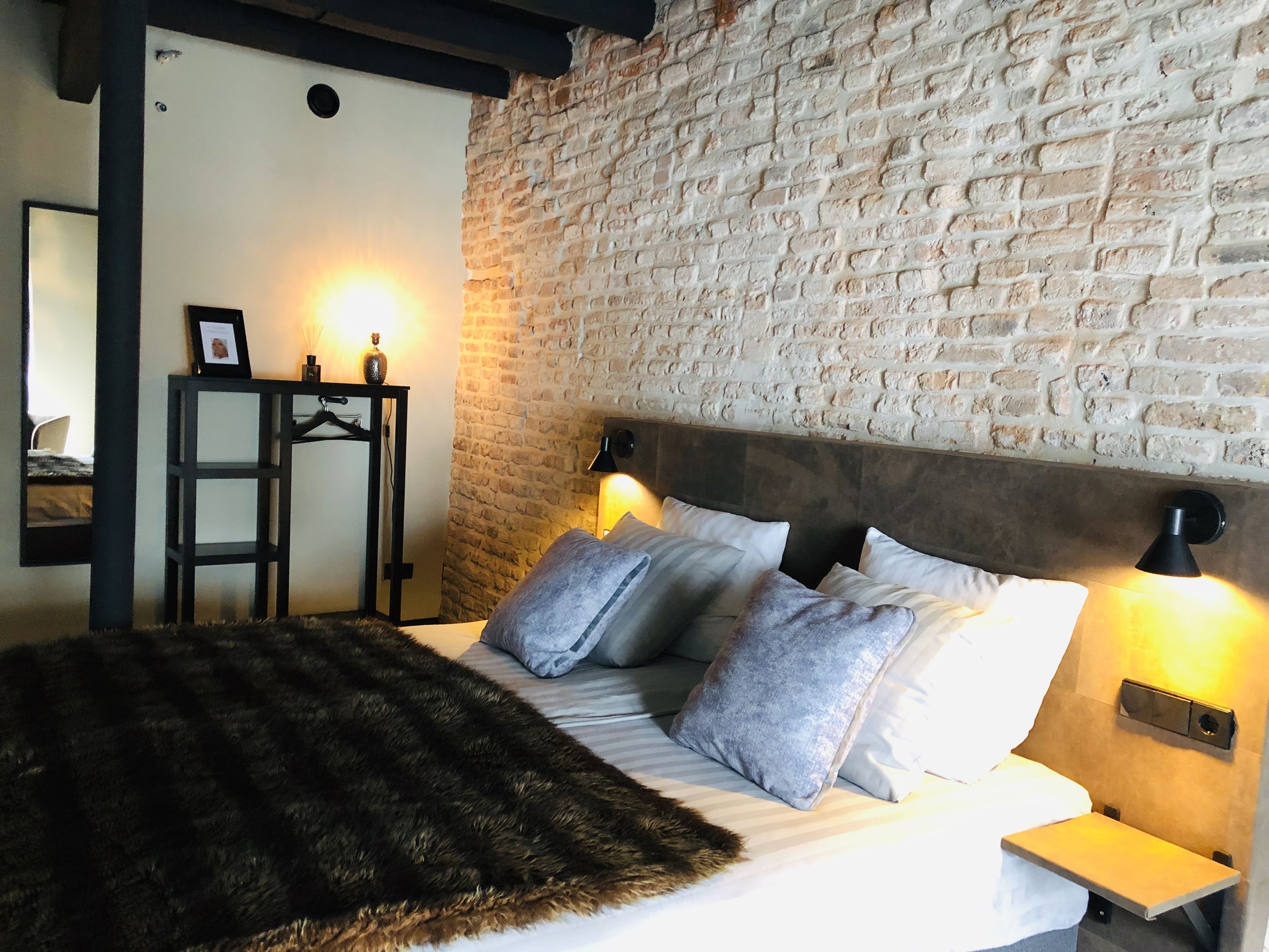 A bedroom with a brick wall, a bed with grey and white pillows, a dark furry throw, and two bedside lamps.