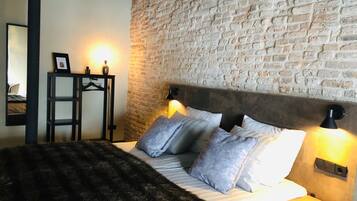 A bedroom with a brick wall, a bed with grey and white pillows, a dark furry throw, and two bedside lamps.