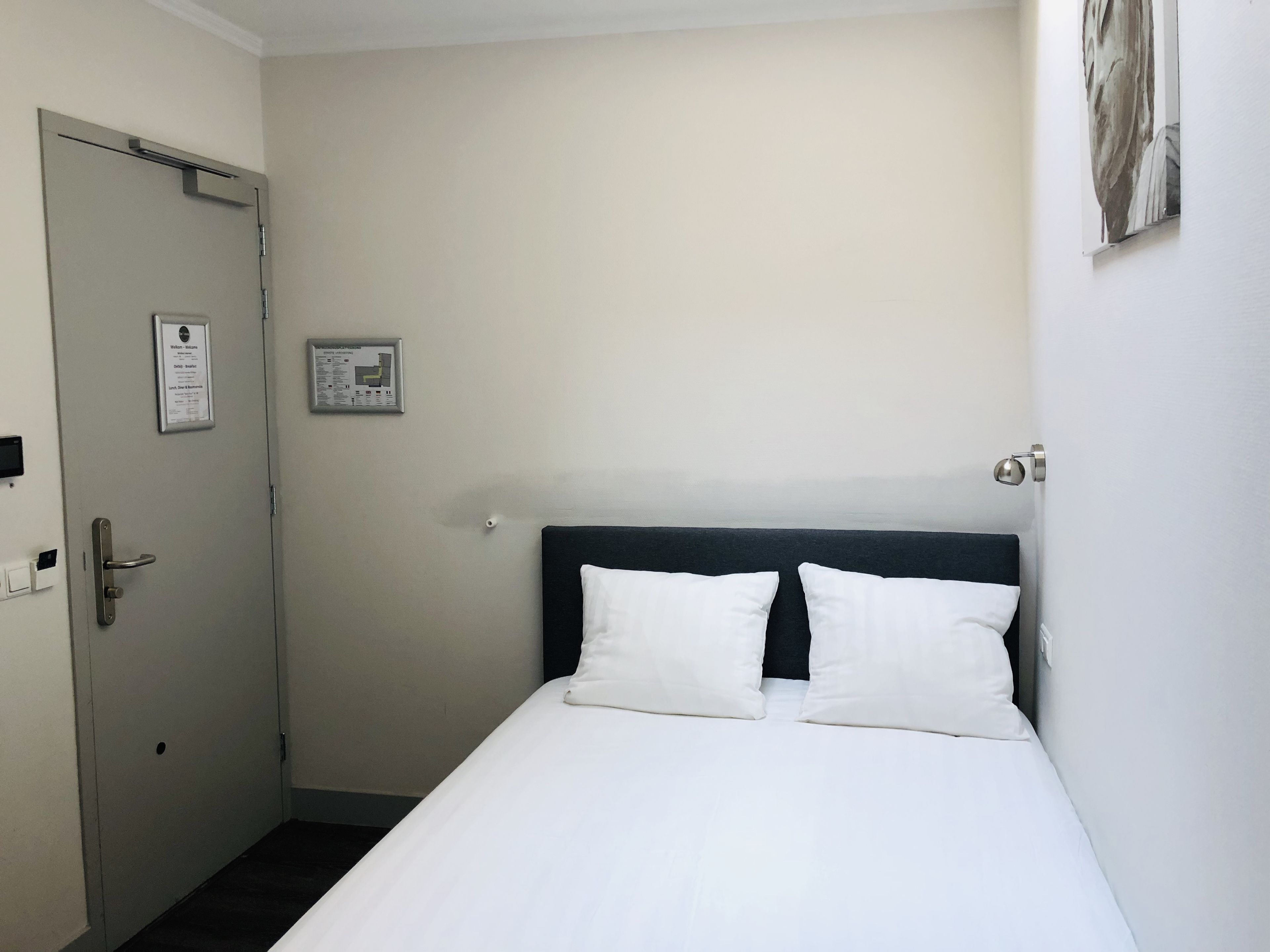 A hotel room with a bed, a door, a wall-mounted light, and a framed certificate.