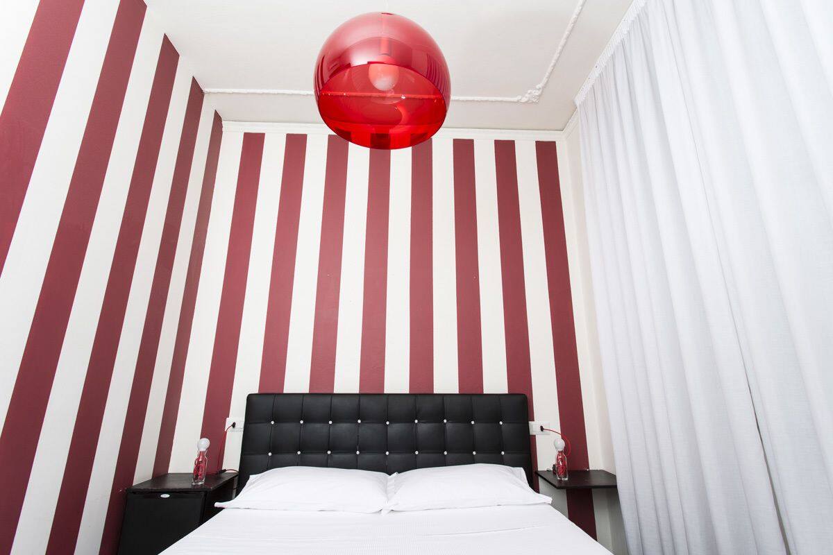 Habitación de hotel con pared a rayas rojas y blancas, cabecero negro acolchado, cama blanca y lámpara esférica roja.