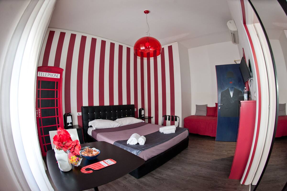 Una habitación de hotel con una cama, un escritorio con un florero y un frutero, un teléfono rojo y una lámpara colgante roja.