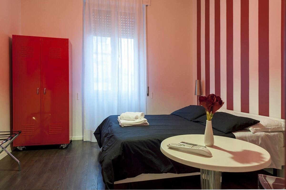 Una habitación de hotel con una cama, un armario rojo, una mesa redonda con un control remoto y un florero con flores.